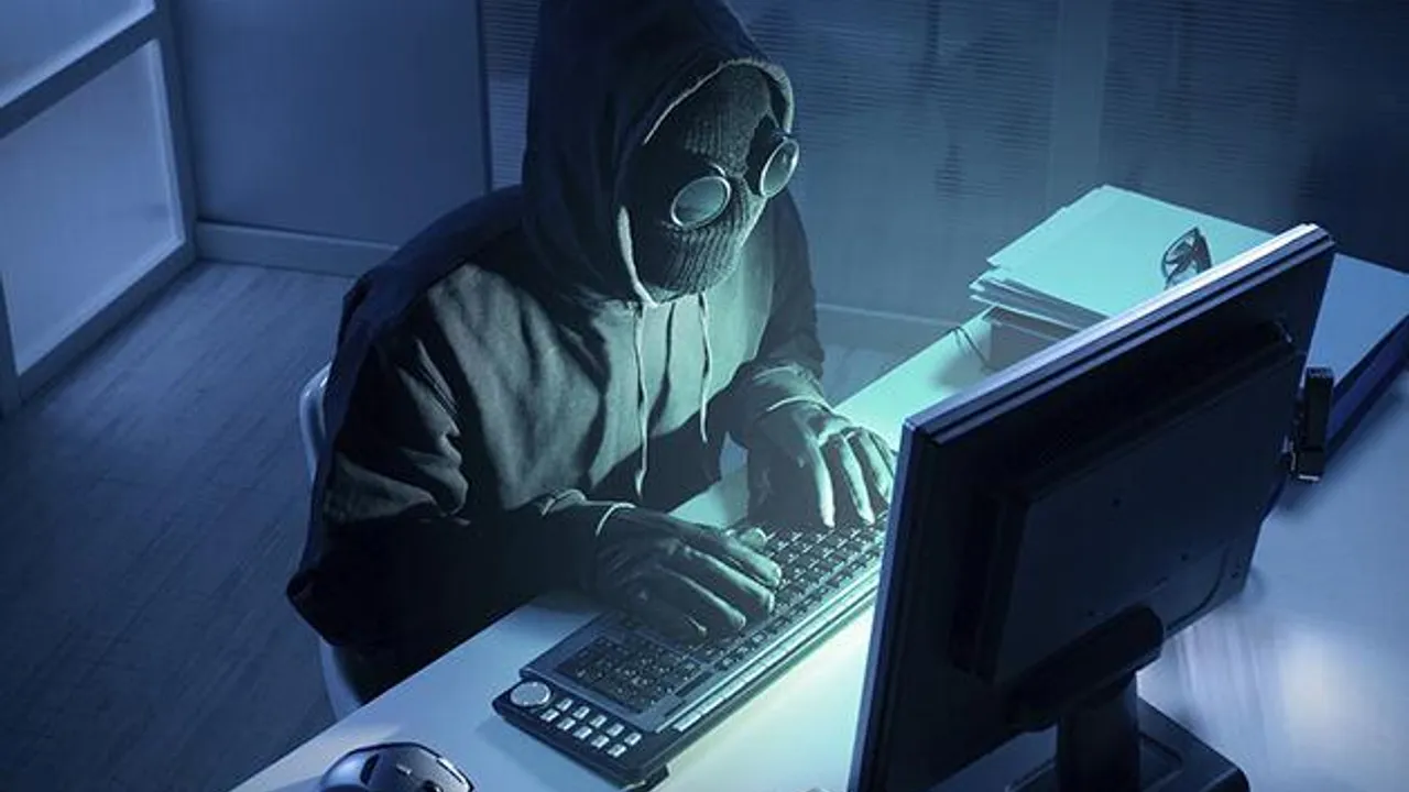 Persona con capucha y pasamontañas teclea frente a un monitor de computadora en una habitación oscura
