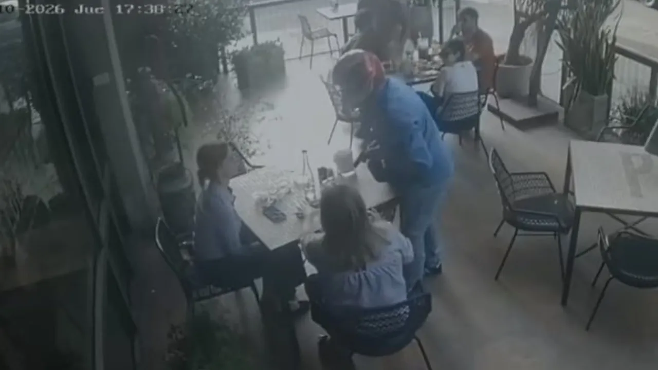 Cámara de seguridad muestra a un asaltante con casco y arma apuntando a clientes sentados en la terraza de un café.