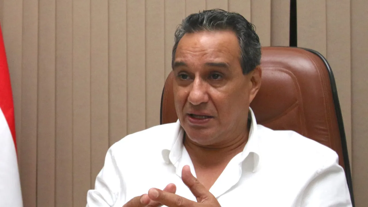 Hombre de camisa blanca gesticula con las manos en un despacho con cortinas de fondo