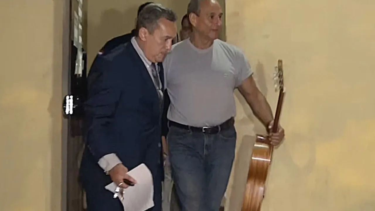 Hugo Javier González camina vistiendo una remera gris y jeans mientras sostiene una guitarra clásica junto a un hombre de traje.