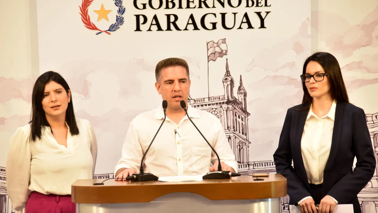 Un hombre habla en un atril con micrófonos acompañado de dos mujeres en una conferencia oficial.