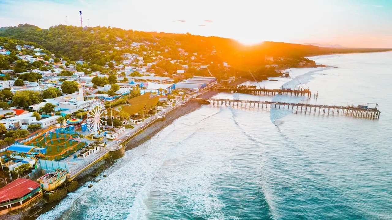 Vista aérea costera al atardecer con muelle sobre el mar, parque de diversiones con noria y poblado en la ladera.