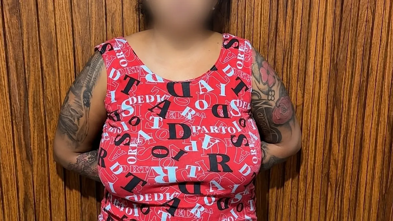 Mujer con camiseta roja estampada y tatuajes en los brazos con las manos en la espalda y rostro difuminado.