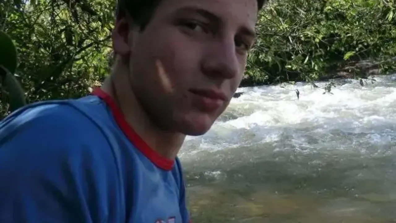 Joven con remera azul mirando de cerca a la cámara al borde de un río con vegetación boscosa de fondo.