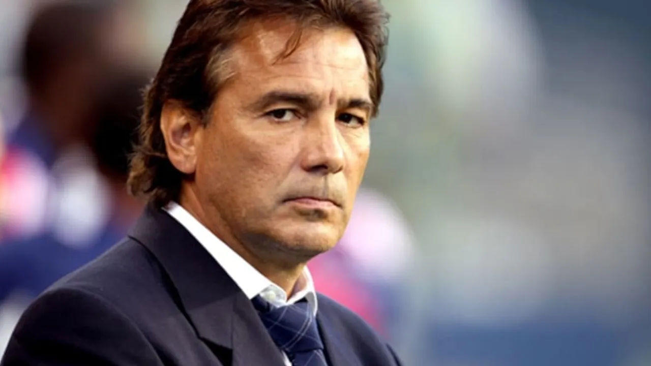 Hombre de cabello castaño y traje oscuro con corbata azul mirando con expresión seria durante un partido.