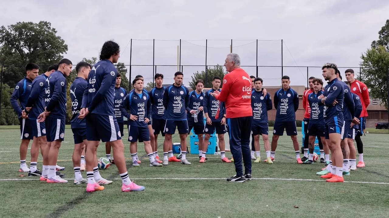 Futbolistas en ronda escuchando las instrucciones de un entrenador con campera roja en una cancha de césped.