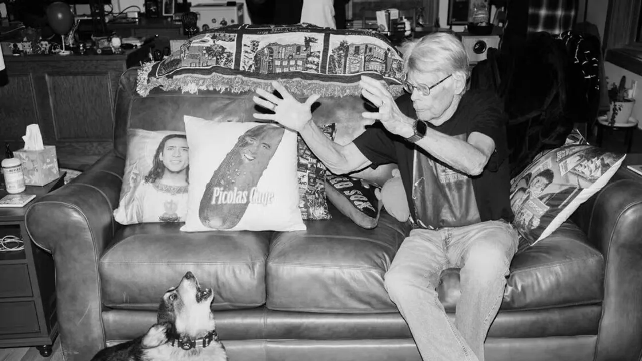 Stephen King gesticula sentado en un sofá junto a un perro corgi que ladra, en una sala con almohadones decorados.