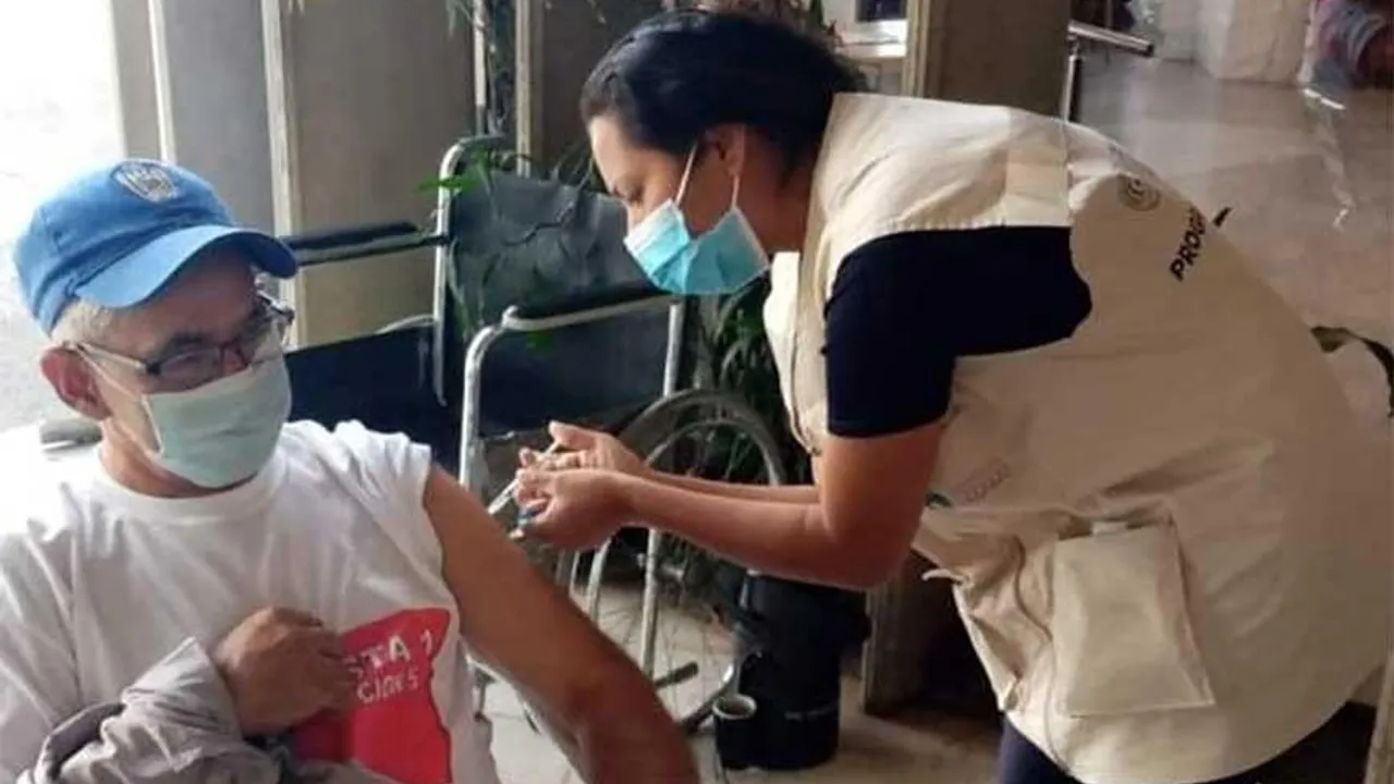 Profesional de salud con mascarilla aplica una inyección en el brazo a un hombre sentado en silla de ruedas.