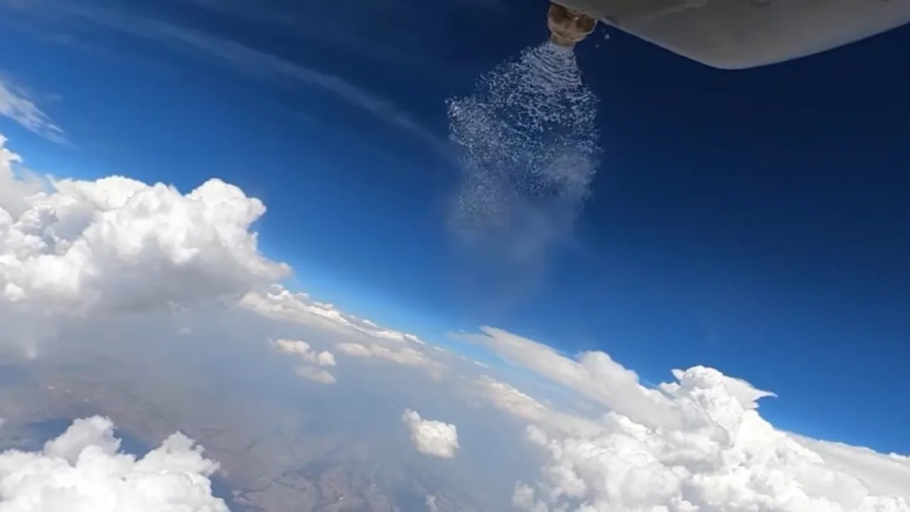 Vista desde el aire de un dispositivo expulsando partículas sobre formaciones de nubes blancas bajo un cielo azul brillante.