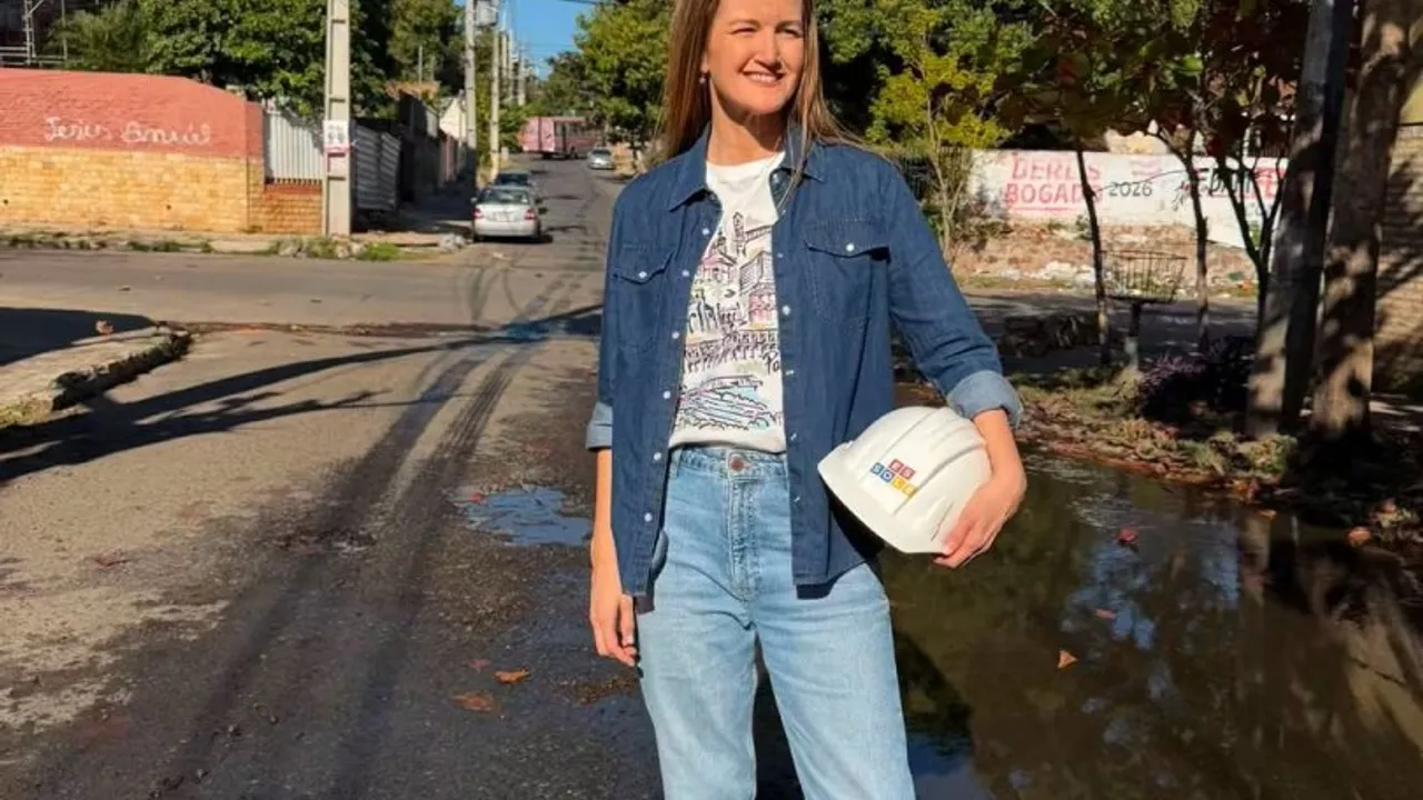 Mujer con camisa de jeans sonríe de pie en una calle asfaltada sosteniendo un casco blanco de seguridad de obra.