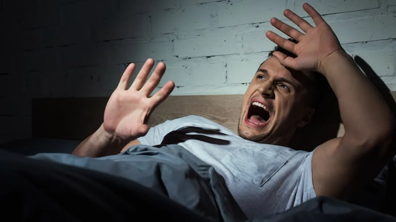 Hombre en la cama despierta asustado con la boca abierta y las manos levantadas en un cuarto oscuro.