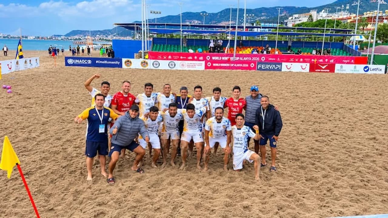 Jugadores y cuerpo técnico de fútbol playa posan juntos sobre la arena frente a las gradas del estadio.