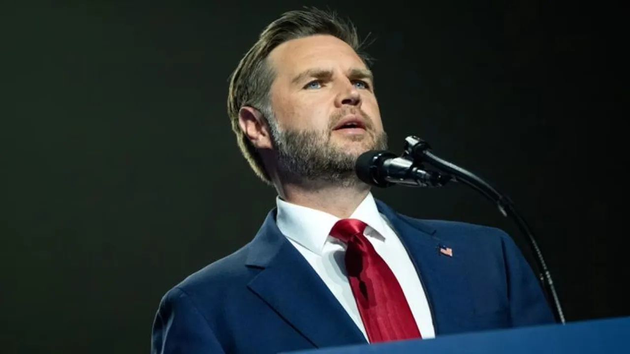 J. D. Vance habla ante un micrófono con traje oscuro y corbata roja en un escenario iluminado.