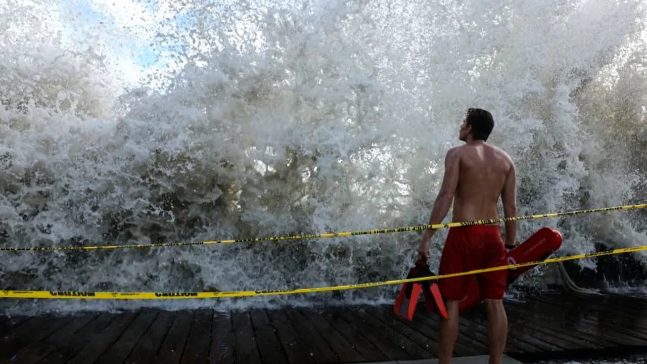 Salvavidas en la costa observa una ola gigante romper tras una cinta de precaución amarilla en el mar.