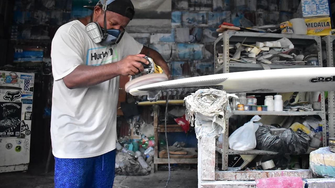 Hombre con mascarilla y gorra lija una tabla de surf con una pulidora eléctrica dentro de un taller artesanal.