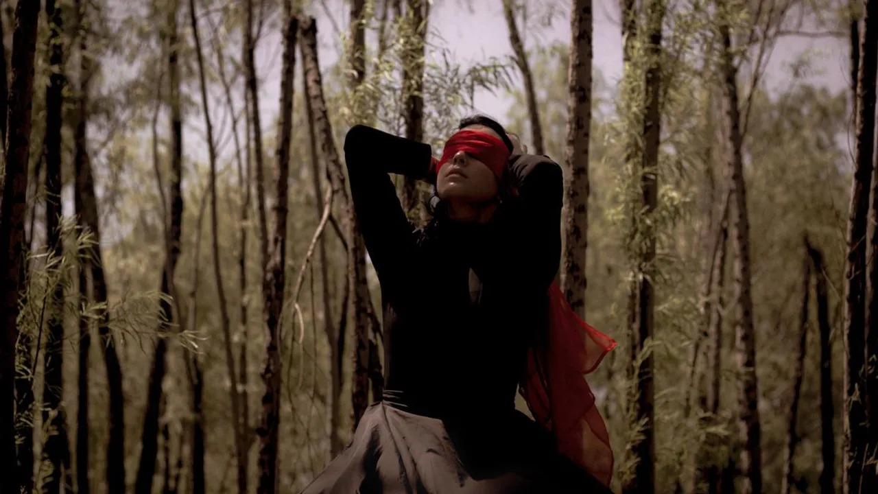 Persona vestida de negro y con los ojos vendados de rojo entre árboles de un bosque, con los brazos en la cabeza.