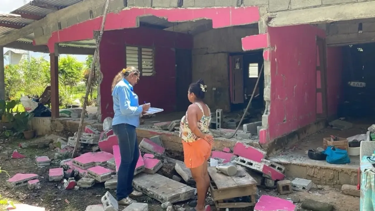 Dos mujeres dialogan frente a una vivienda de color rosa con paredes derrumbadas y escombros esparcidos en el suelo.