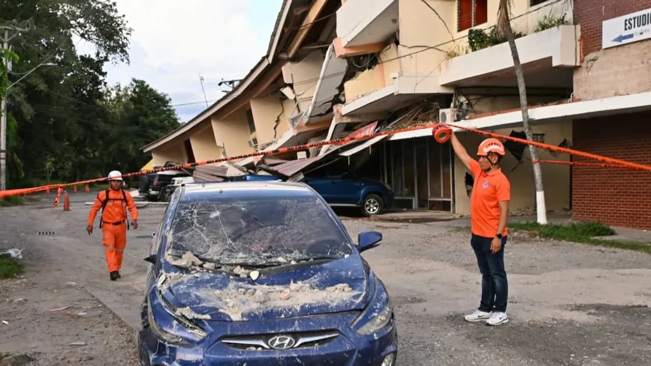Rescatistas con cascos colocan cinta de peligro ante un edificio colapsado y un automóvil cubierto de escombros.