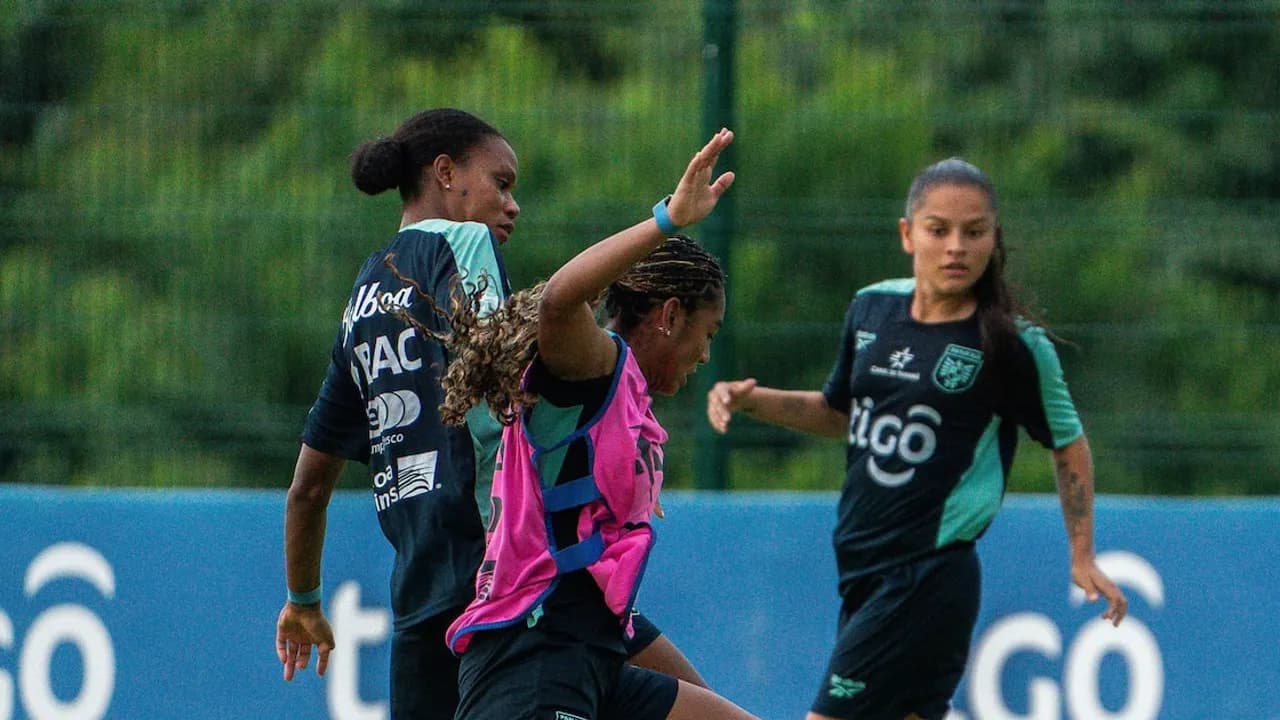 Jugadoras de fútbol entrenan en un campo verde vestidas con indumentaria deportiva oscura y pecheras de práctica.