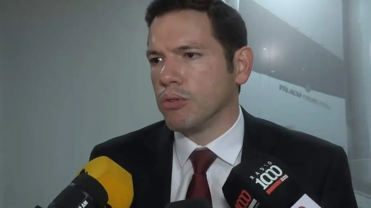 Hombre de traje oscuro y corbata bordó hablando frente a micrófonos de prensa en un pasillo.