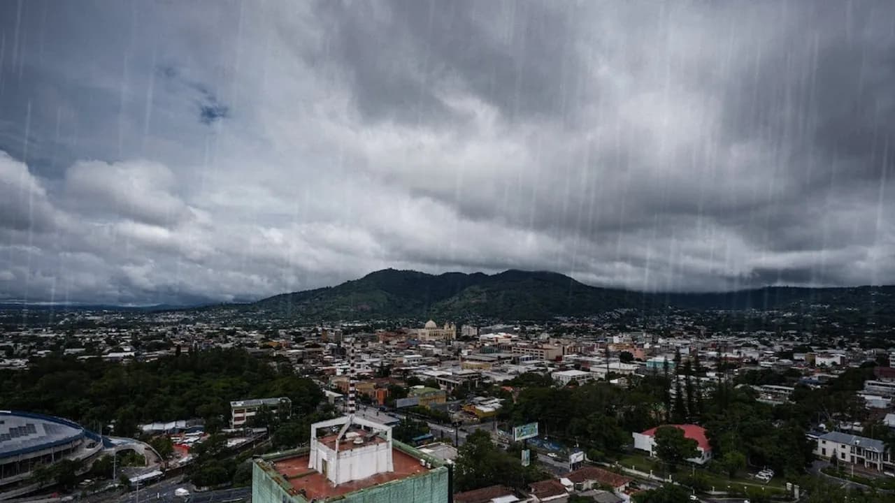 Vista panorámica aérea de la ciudad bajo un cielo cubierto de nubes densas y cortinas de lluvia en las montañas.