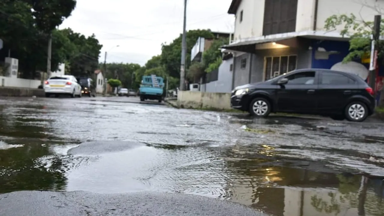 Calle urbana anegada con raudales y baches de agua mientras varios automóviles circulan con precaución.