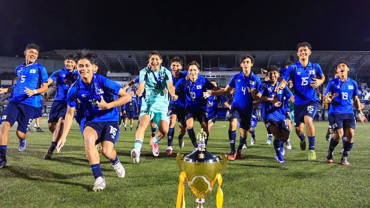 Futbolistas juveniles vestidos de azul festejan corriendo en la cancha detrás de un trofeo dorado con cinta amarilla.