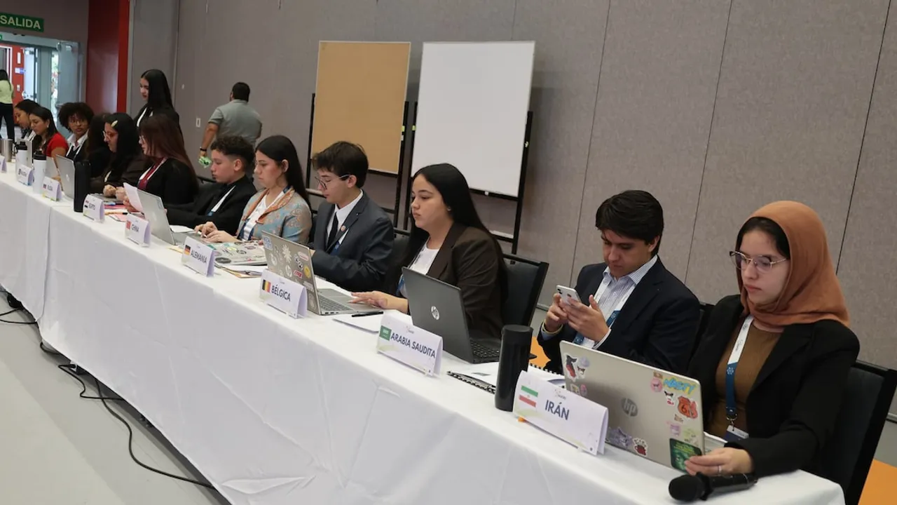 Jóvenes sentados frente a computadoras portátiles en una mesa con carteles de países durante una simulación diplomática.