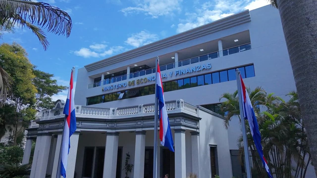 Fachada del Ministerio de Economía y Finanzas con banderas paraguayas en el acceso principal y cielo despejado.