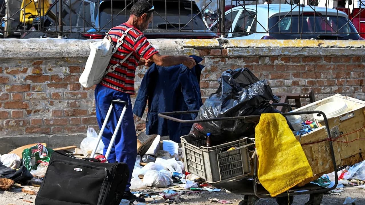 Hombre de espaldas revisando una prenda frente a un carro cargado de bolsas y cajas en una calle.