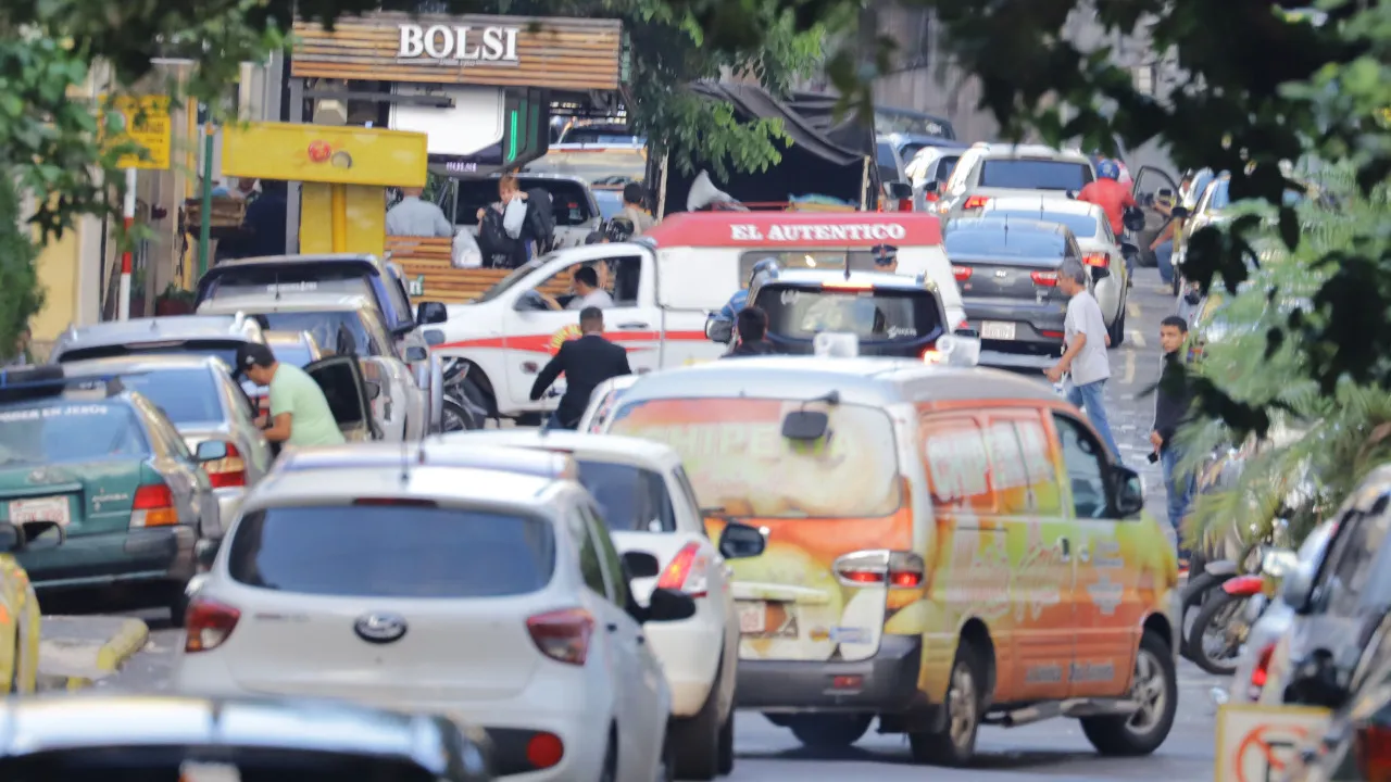 Tránsito vehicular congestionado en una calle céntrica con autos, furgones y peatones circulando