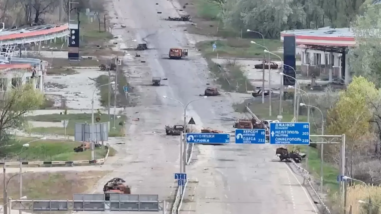 Carretera desolada con vehículos destruidos, carteles viales y restos de daños por combates en una zona urbana.