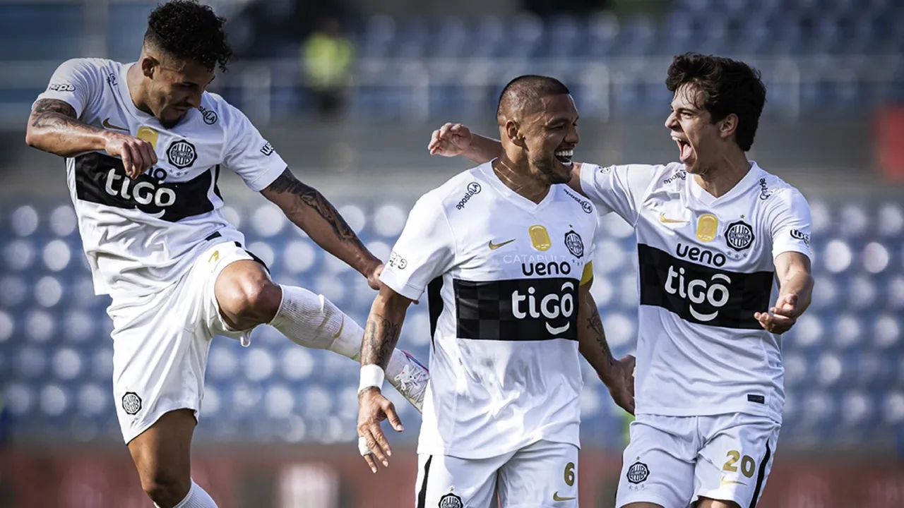 Tres futbolistas con camiseta blanca y franja negra festejan con sonrisas y brazos abiertos en la cancha.