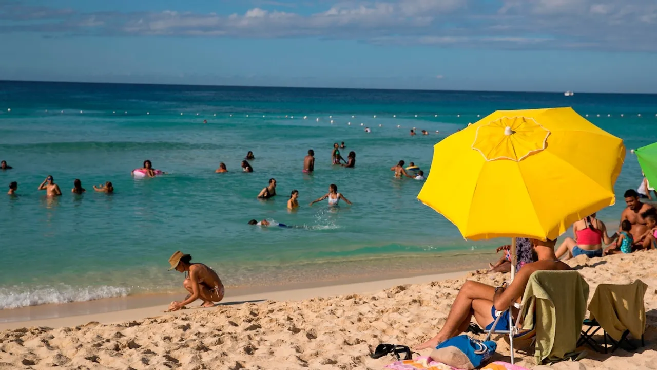 Bañistas disfrutando en el mar turquesa junto a una sombrilla amarilla instalada sobre la arena de una playa caribeña.