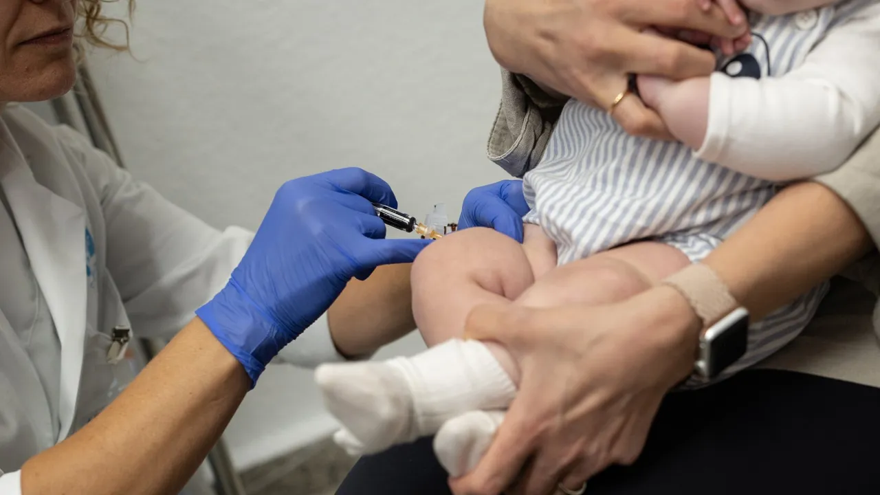 Profesional de blanco con guantes azules aplica una inyección en la pierna de un bebé en brazos de un adulto.