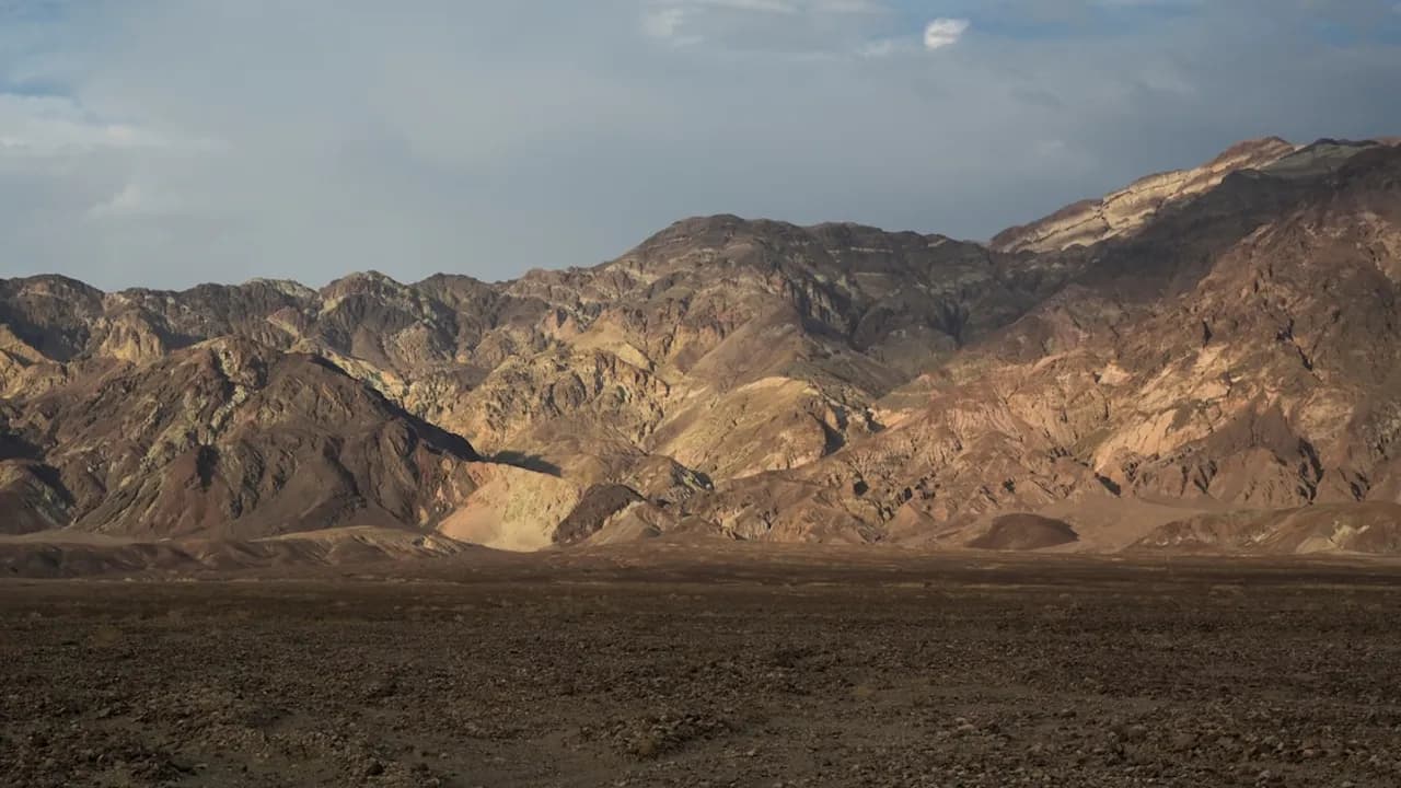 Cadena montañosa árida y rocosa con tonalidades marrones y ocres bajo un cielo parcialmente nublado en una zona desértica.