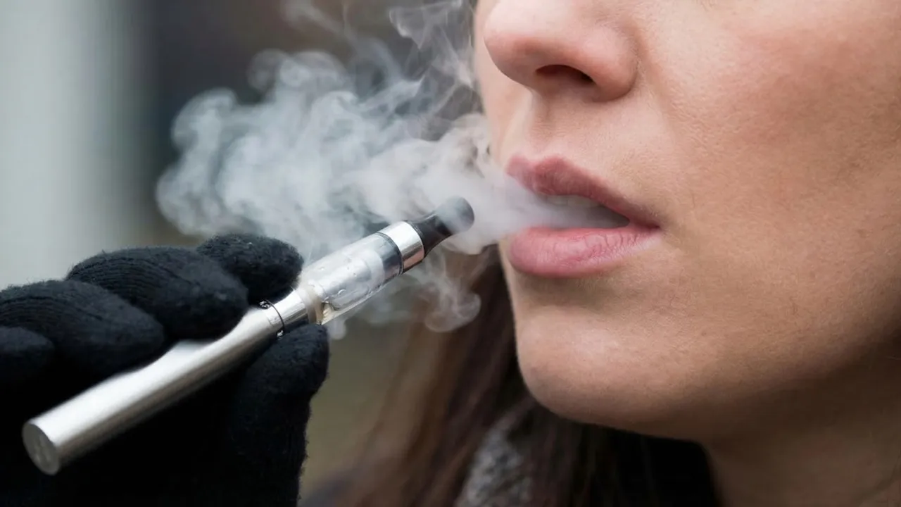 Primer plano de una persona exhalando una densa nube de humo mientras sostiene un cigarrillo electrónico metálico.