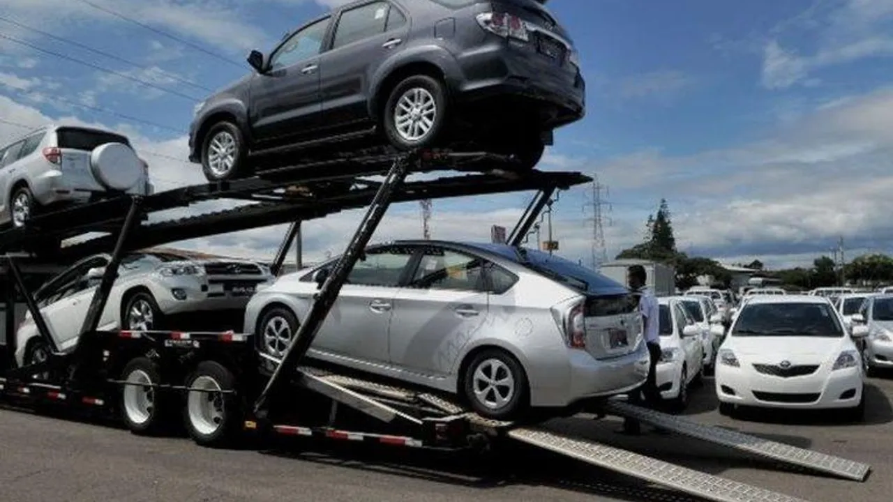 Camión cigüeña de dos pisos transporta automóviles e híbridos en una playa de estacionamiento al aire libre.