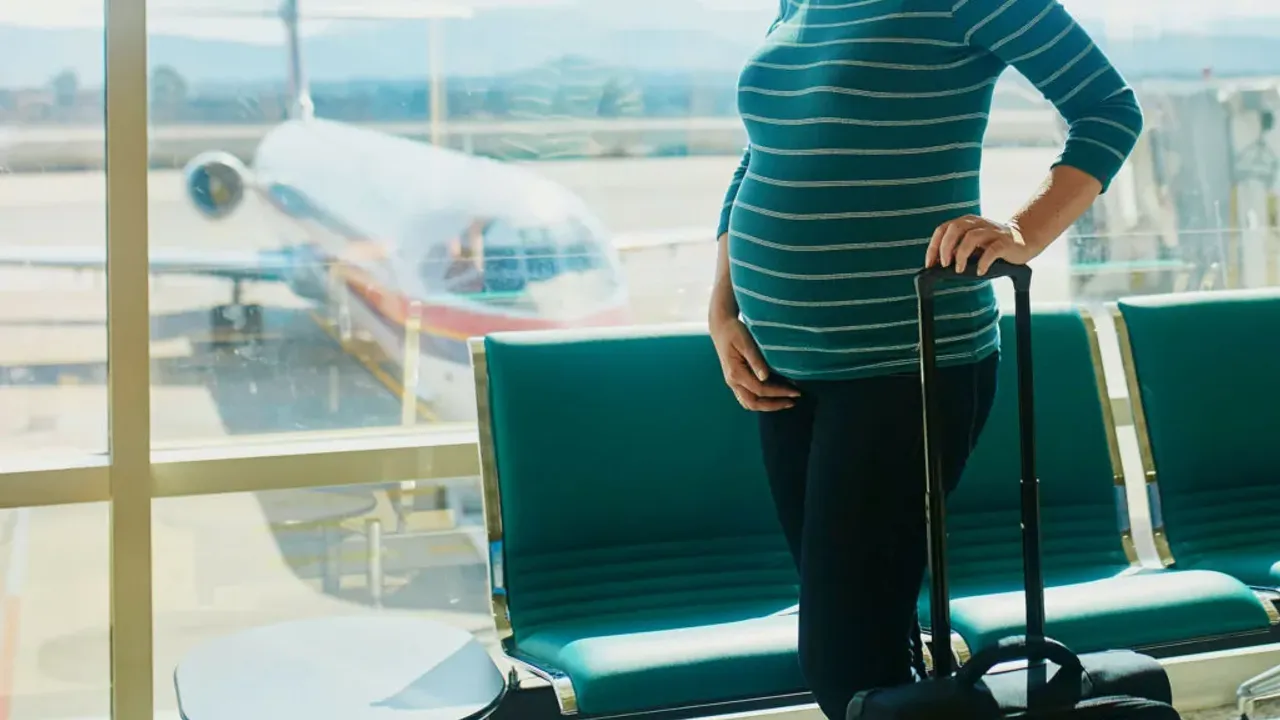 Mujer embarazada de pie junto a su valija en la sala de espera de un aeropuerto frente a un avión.
