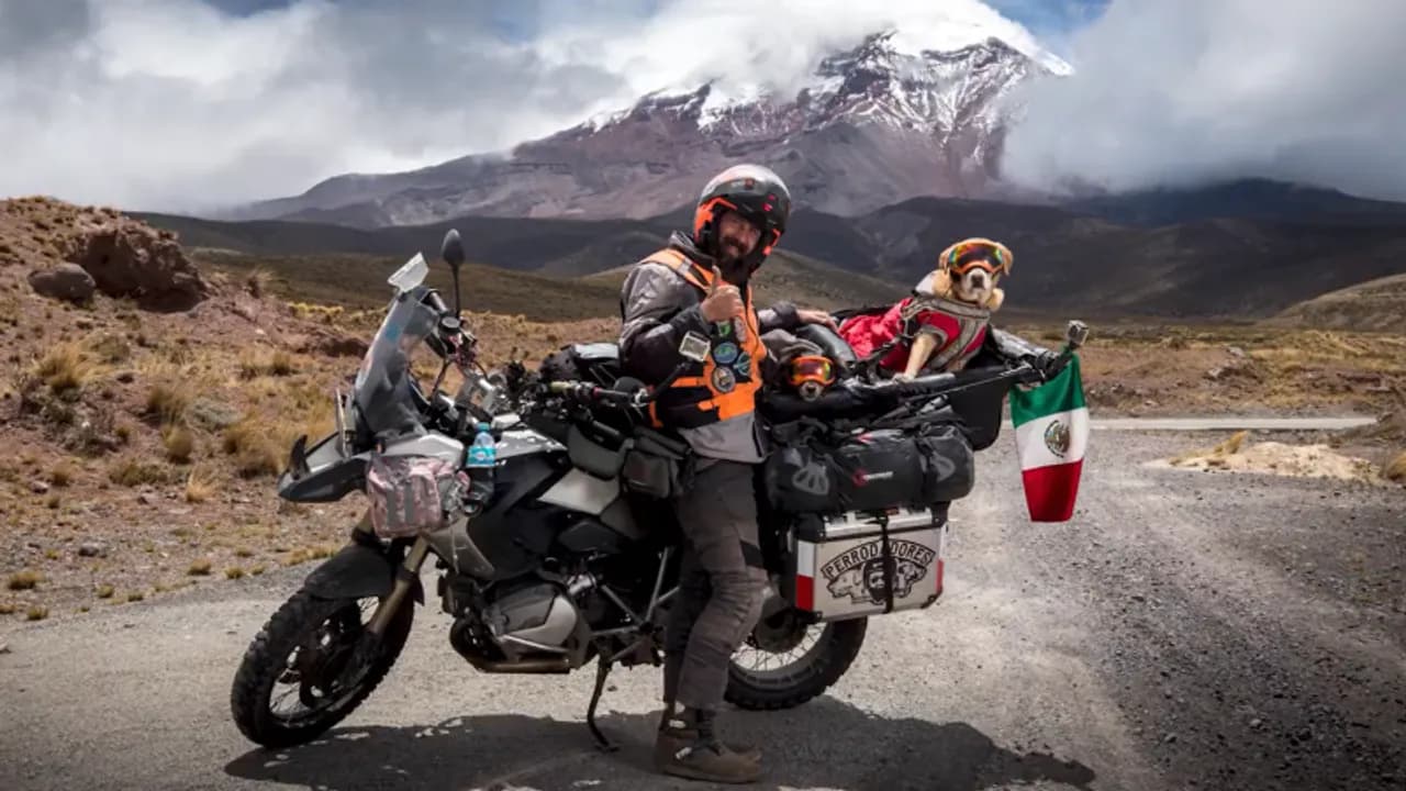 Hombre con casco y motocicleta equipada posa junto a un perro con antiparras en un camino de montaña.
