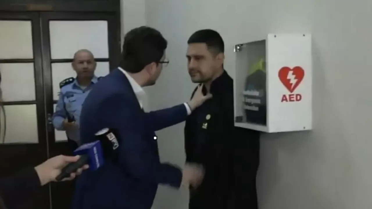 Un hombre de traje azul encara contra la pared a otra persona en un pasillo junto a un desfibrilador.