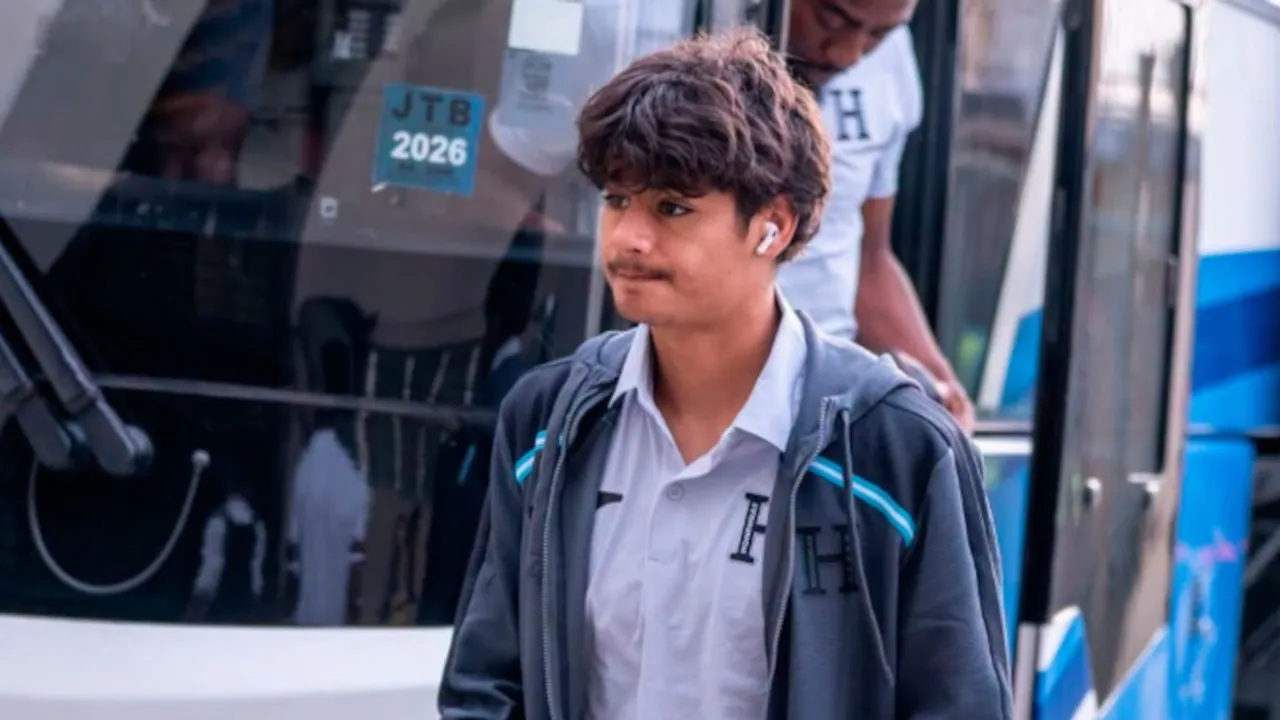 Futbolista joven con campera deportiva gris y auriculares camina frente a un autobús de transporte.