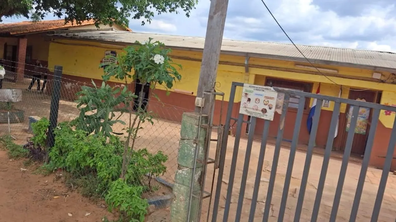 Fachada de la escuela de Limpio con portón de rejas grises y muralla amarilla bajo árboles.