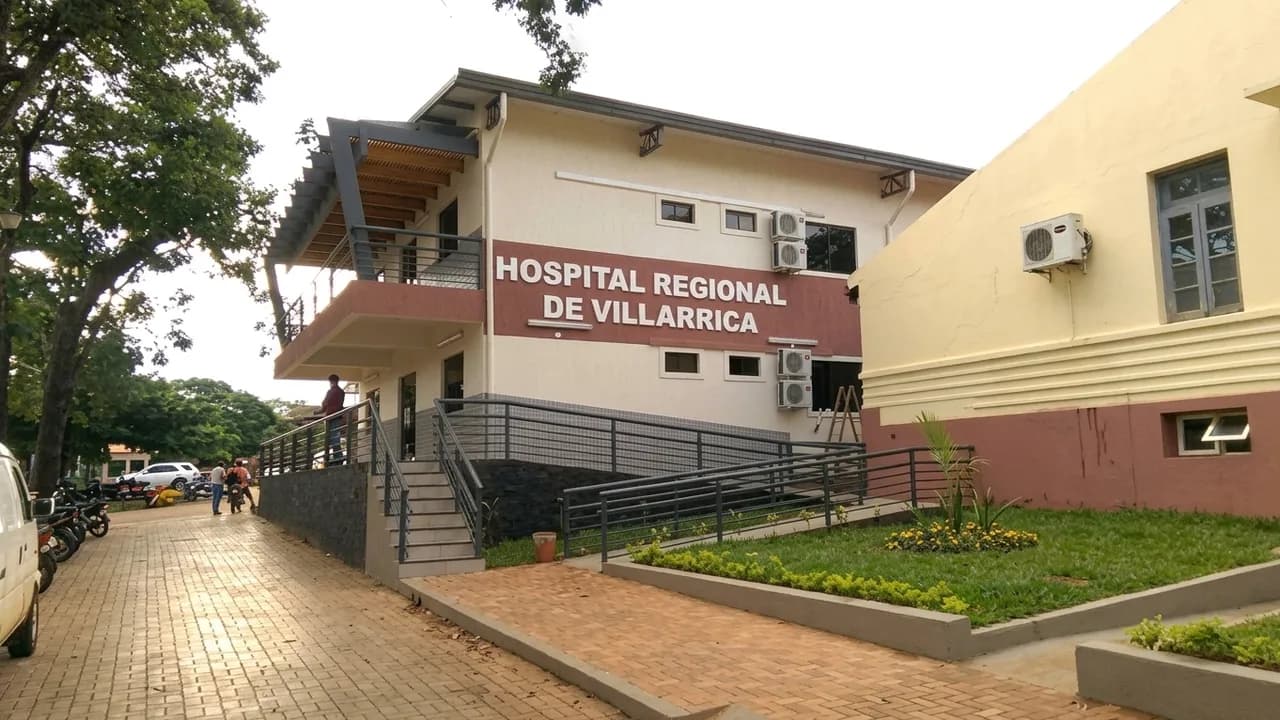 Fachada exterior y rampa de acceso del Hospital Regional de Villarrica con vereda empedrada y árboles.