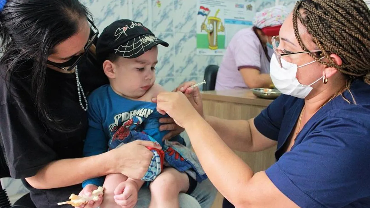 Enfermera con tapabocas aplica una inyección en el brazo de un niño pequeño sostenido por un adulto en un consultorio.