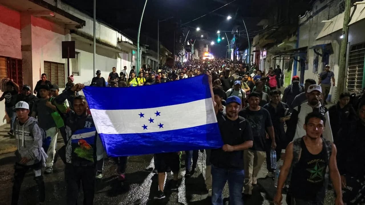 Grupo de migrantes camina de noche por una calle urbana portando una gran bandera de Honduras.
