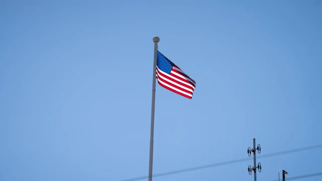 Bandera de Estados Unidos flameando en un mástil blanco bajo un cielo despejado y sin nubes.