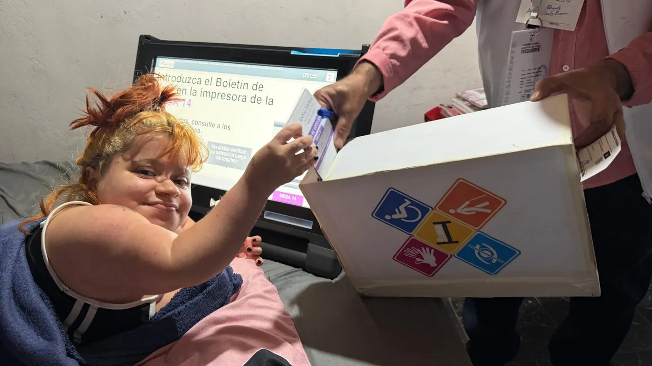 Mujer en cama introduce su boletín en la urna electoral sostenida por un funcionario junto a una máquina de votación