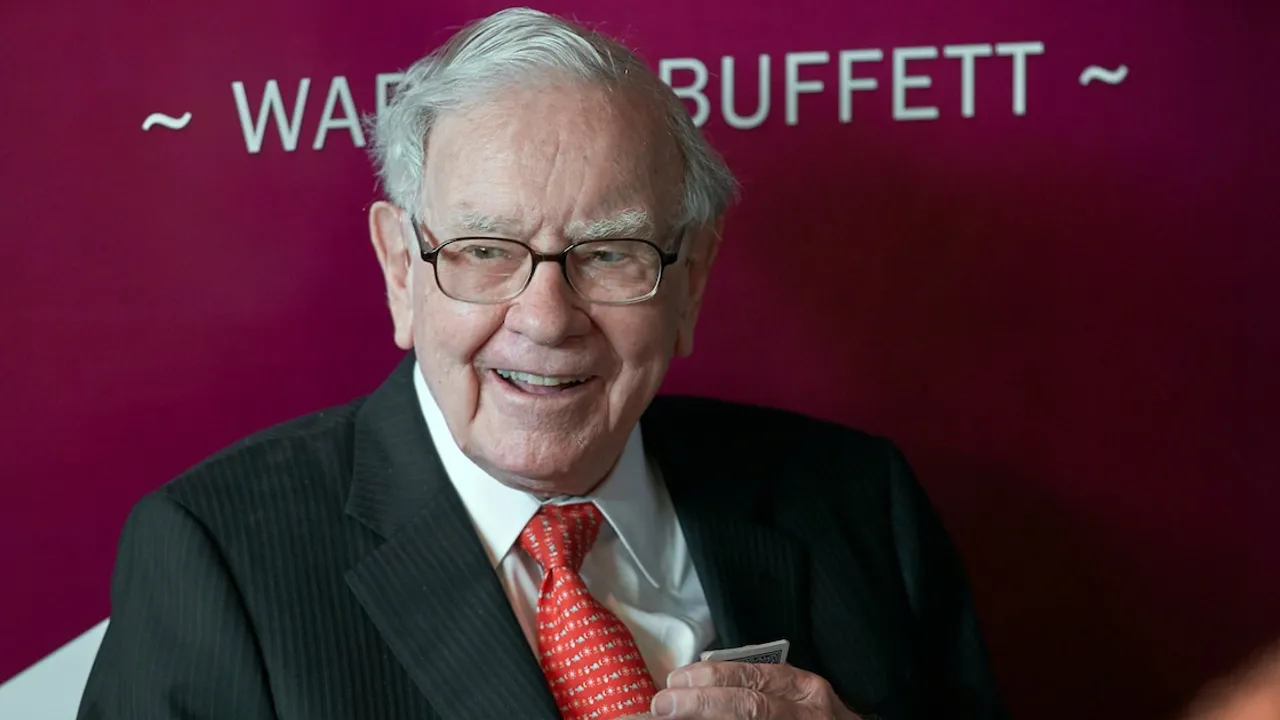 Warren Buffett con traje oscuro, camisa blanca, corbata roja y anteojos, sonriendo frente a un fondo bordó.