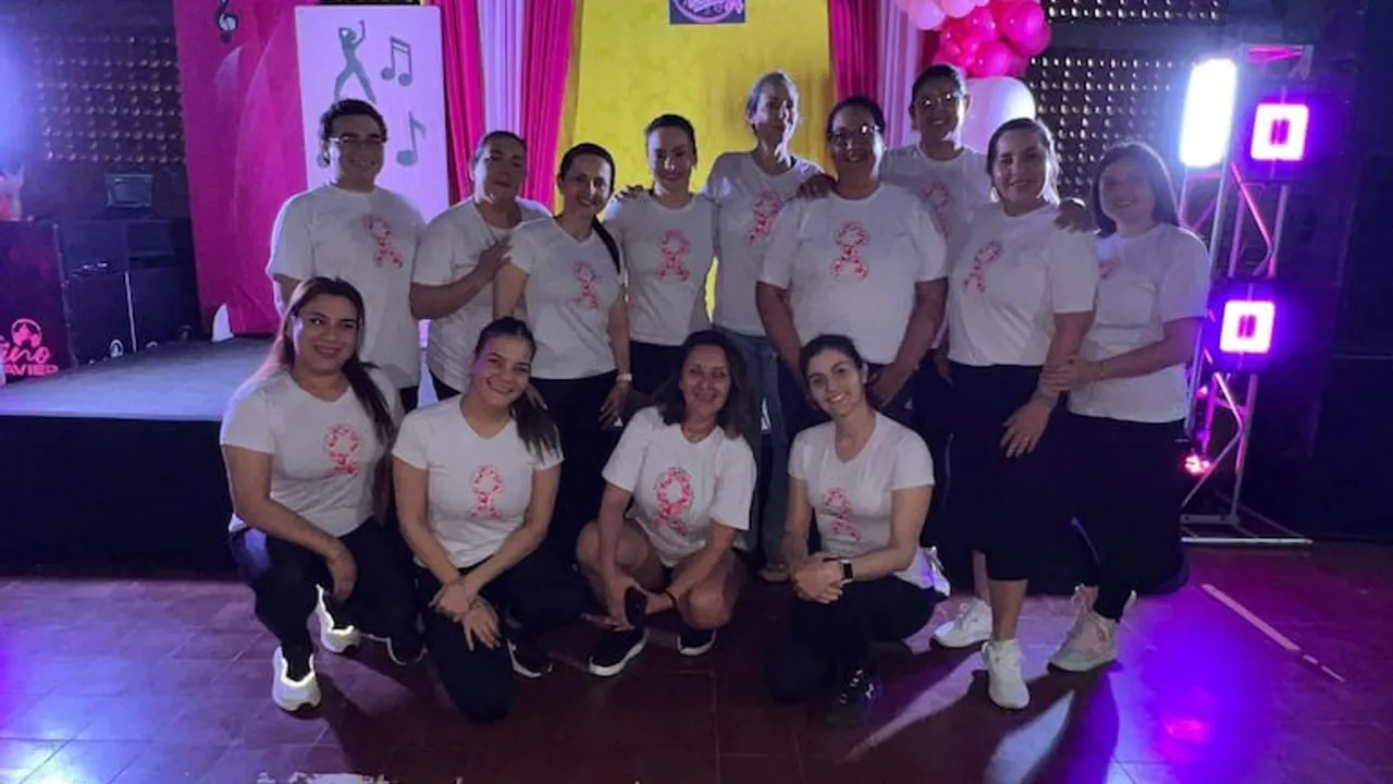 Grupo de mujeres posando con remeras blancas estampadas con un lazo rosa durante un evento de zumba solidario.